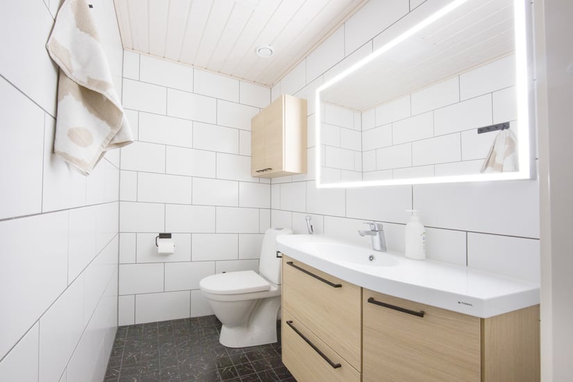 Moderni kylpyhuone isolla valopeilillä, vaaleilla kalusteilla sekä valkoisella seinälaatalla jossa tiililadonta.